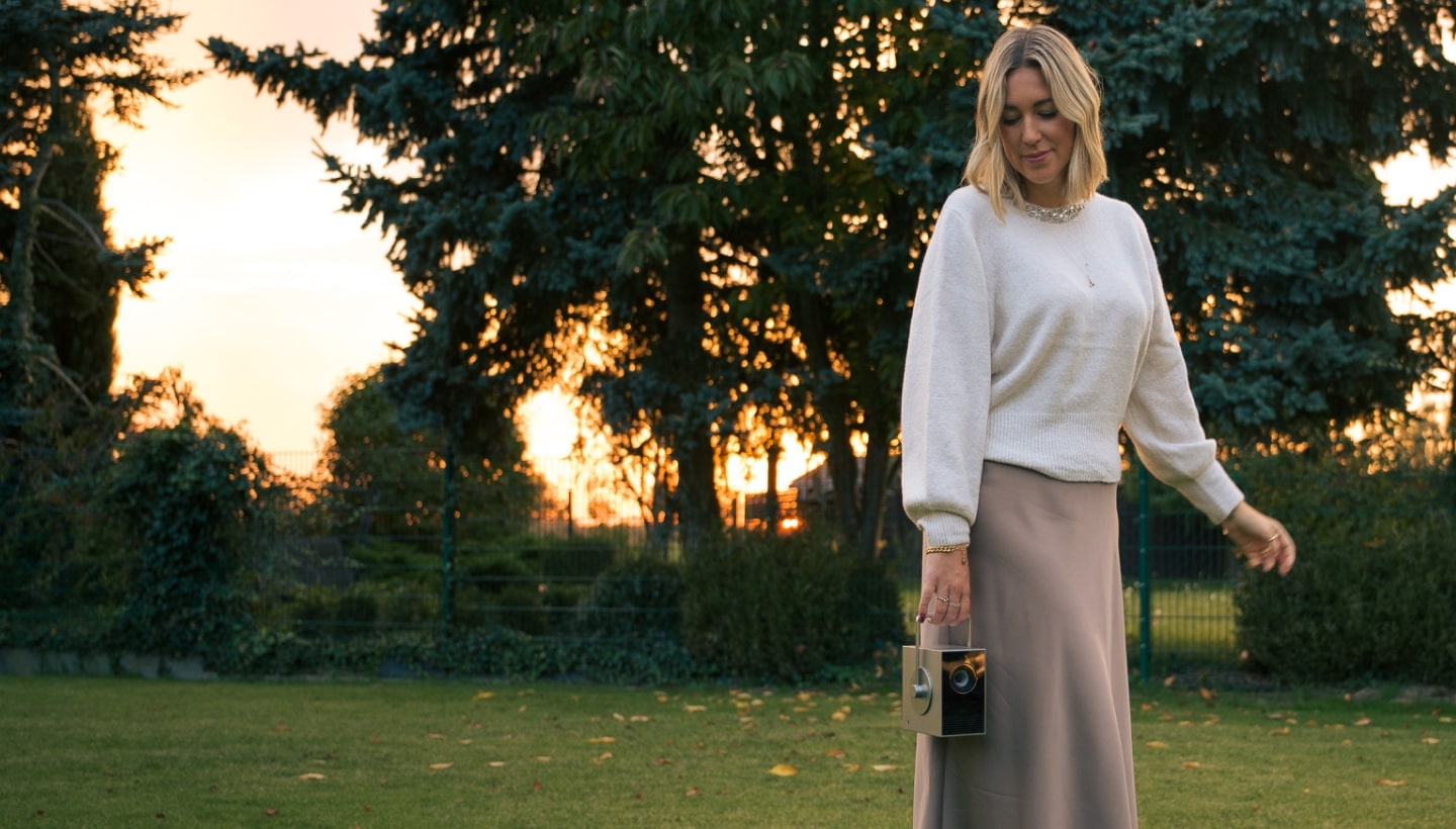 Une femme marche à l'extérieur au coucher du soleil, tenant un projecteur LG CineBeam. Le jardin est entouré d'une végétation luxuriante.