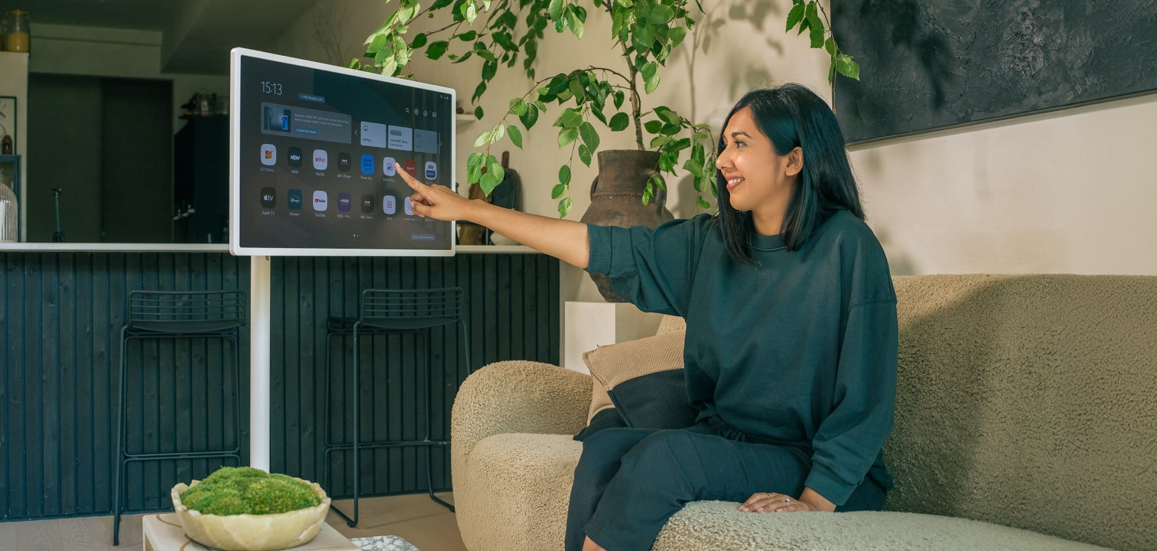 Salon avec une femme interagissant avec l’écran d’un LG StanbyME, assise sur un canapé confortable entourée de plantes.