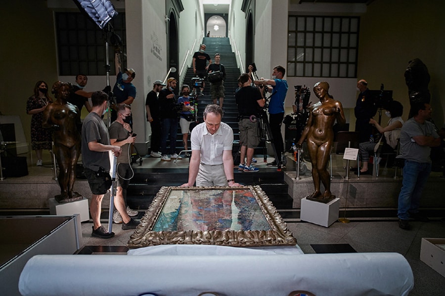 A man looks down at a framed artwork as a film team sets up behind him.