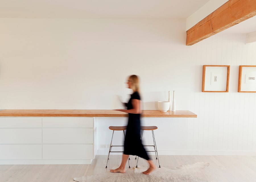 A women in walking motion alongside a minimalistic island table and kitchen bar stools.