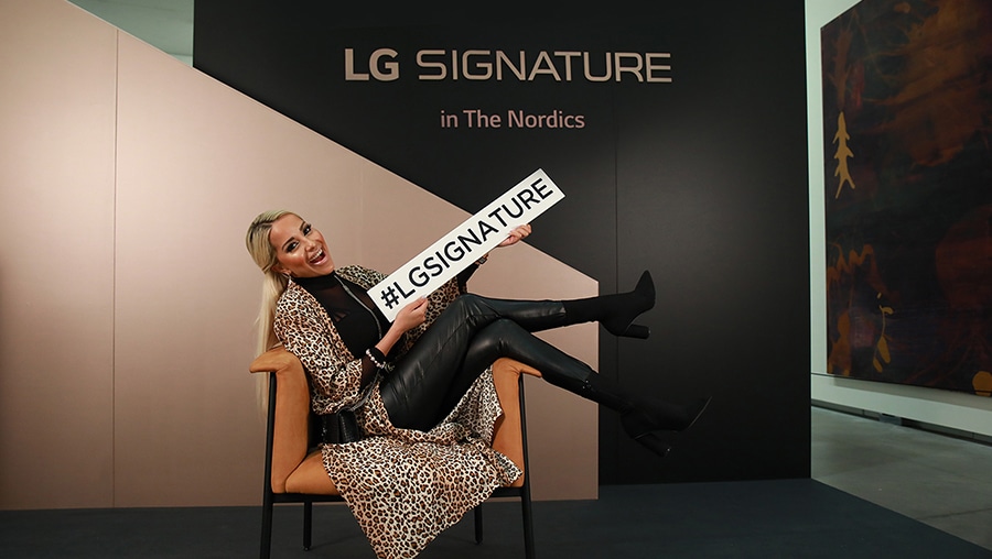 a woman is sitting on a chair and posing while holding lg signature board in front of the photowall at lg signature launching event in nordics