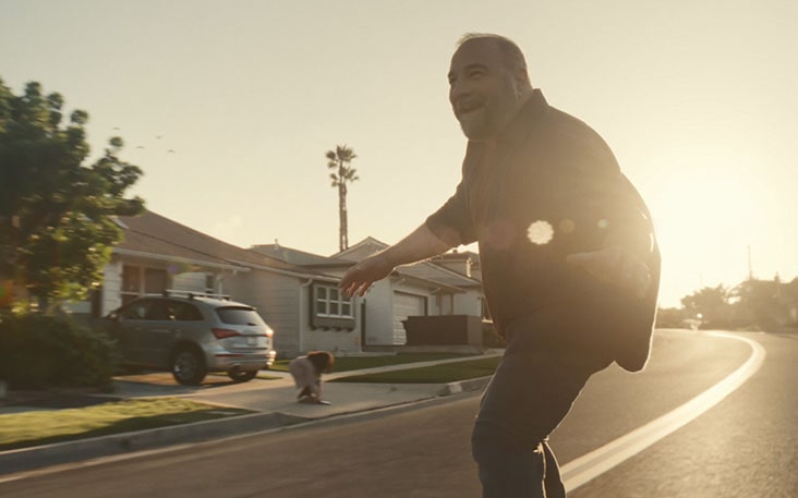 A middle-aged man riding a longboard with bright sunlight shining behind.