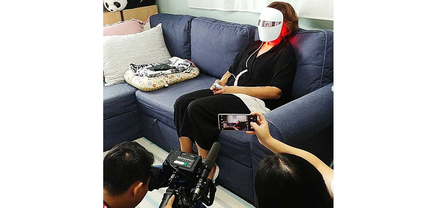 A cameraman shoots a video of a consumer sitting on the couch while wearing the LG Pra.L Derma LED Mask on her face.