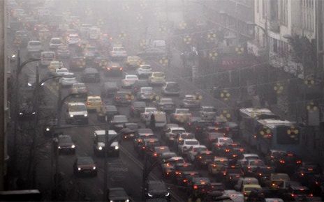 Hundreds of vehicles sit in heavy traffic within a bustling city, while surrounded by heavily polluted air