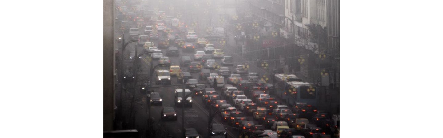 Hundreds of vehicles sit in heavy traffic within a bustling city, while surrounded by heavily polluted air