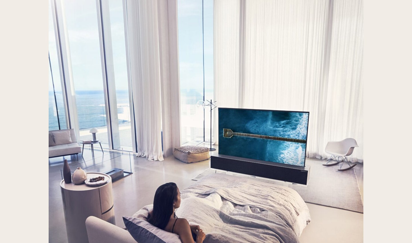 A woman in bed watching an LG SIGNATURE OLED TV R at a bright, beachside mansion.