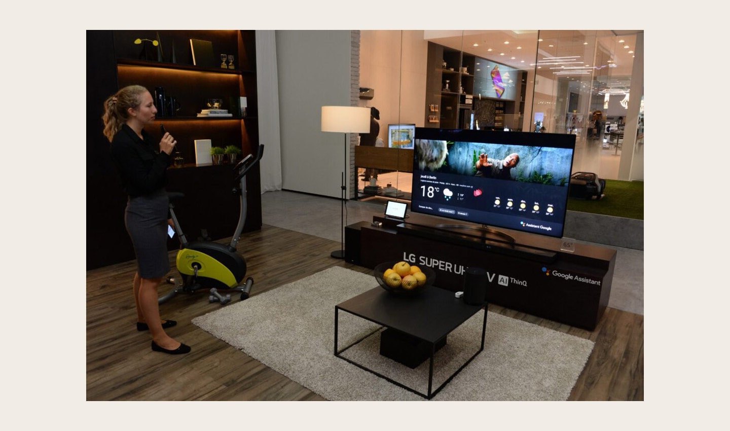 A female attendant holding the Magic Remote stands next to the LG SUPER UHD TV AI ThinQ with Google Assistant to try out the AI voice assistant feature.