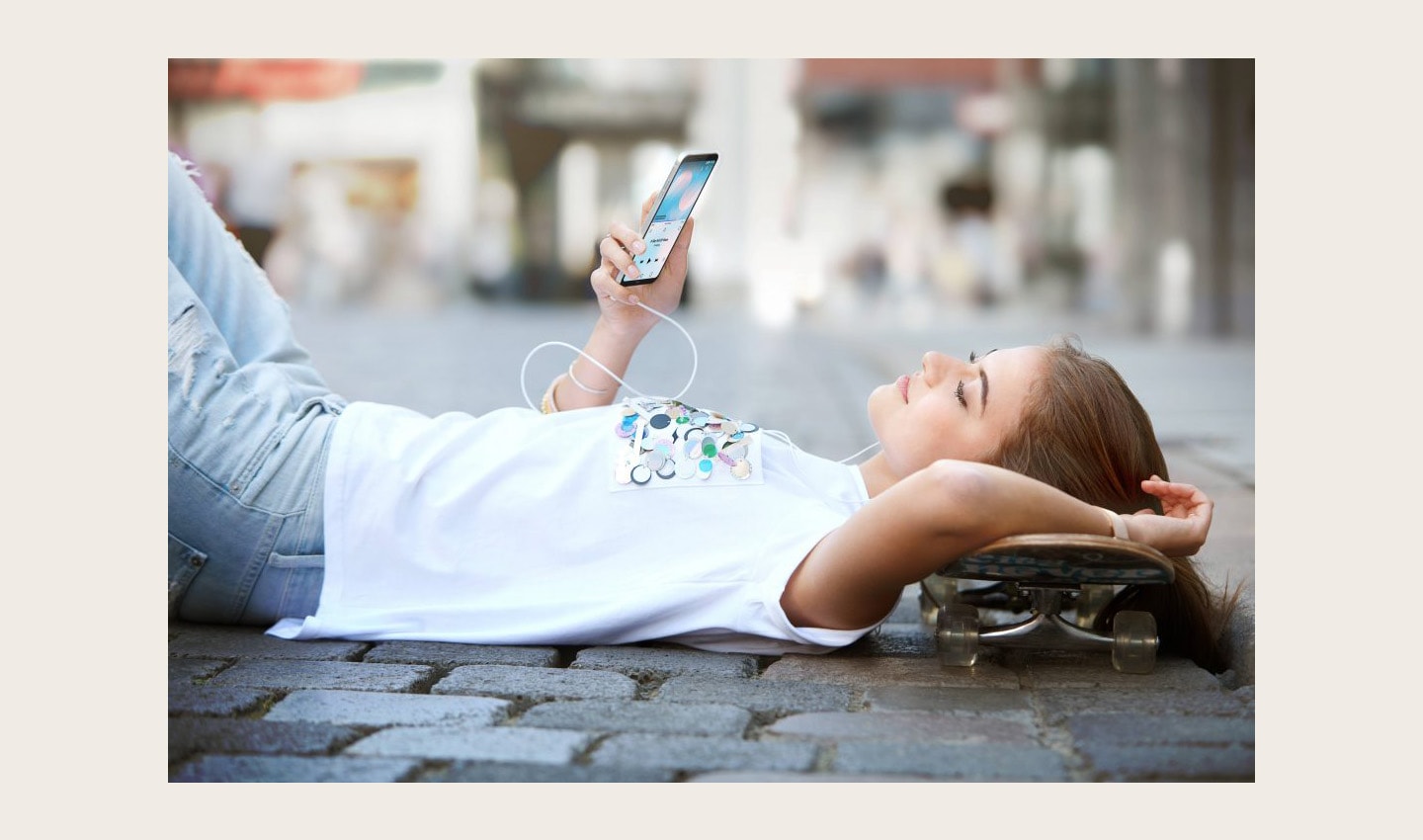 A woman lies on the ground with her skateboard while listening to music on headphones on her LG Q6