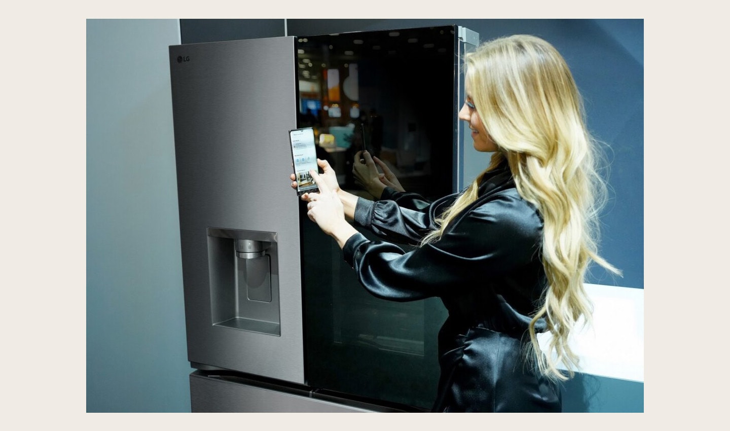 A woman is showing the LG ThinQ app on a smartphone in front of the InstaView fridge at the Kitchen and Bath Industry Show 2023