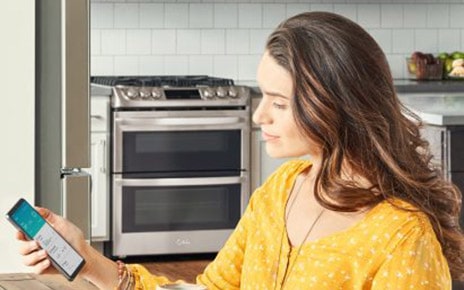Woman sitting in her kitchen looking at a phone displaying the ThinQ™ app screen showing the device status of the washer, refrigerator and range