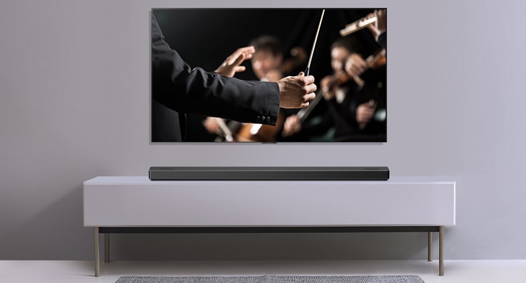 灰色牆上有一個電視，而 LG Soundbar 則在其下方的灰色架子上。電視展示指揮家正指揮樂團。