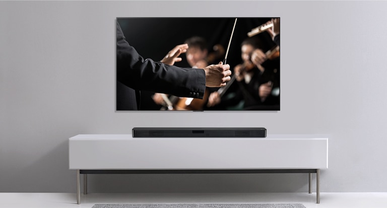 灰色牆上有一個電視，而 LG Soundbar 則在其下方的灰色架子上。電視展示指揮家正指揮樂團。
