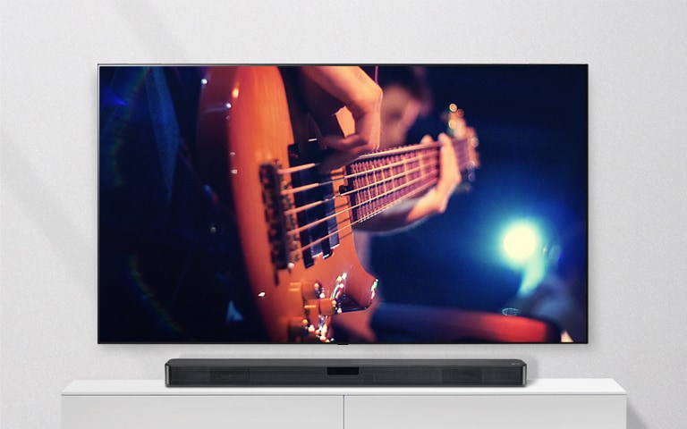 電視貼牆，Sound Bar 置於白色層架上。電視畫面中有位男士正在彈結他。
