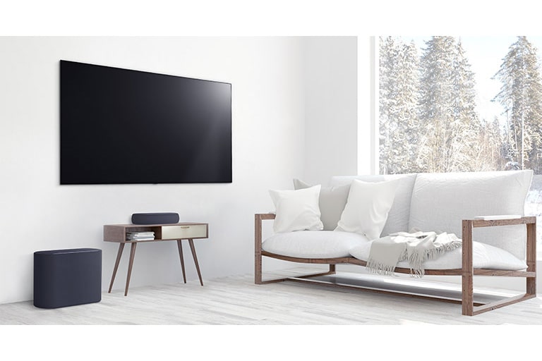 A soundbar is placed on a small tea table,  a subwoofer placed on the left side and TV is placed above a soundbar. The room is overall white.