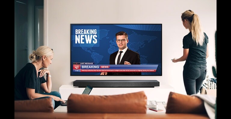 A woman watching the news at its best quality on TV standing up and another sitting in front of her laptop (move the slide)