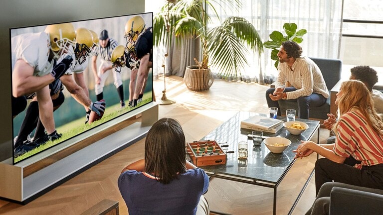 In the living room, a group of friends sitting with a wide viewing angle and watching a football game on TV