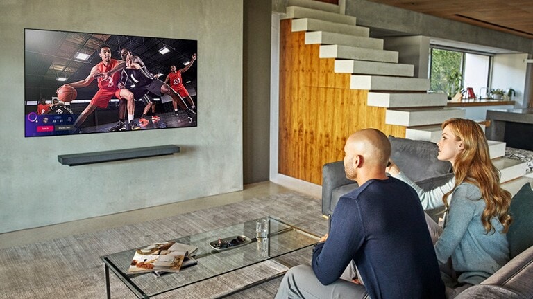 A sports alert of other matches' score pops up at the left bottom of the TV while a couple watching the basketball game on TV