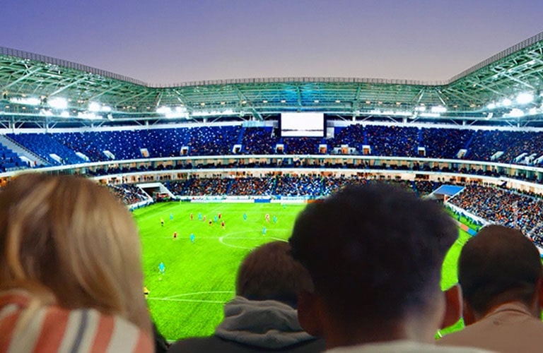 People are watching a soccer game, with a backdrop of the entire stadium, on a large TV