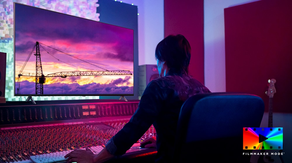 A movie director is looking at a big TV monitor, editing something. The TV screen shows a tower crane in purple sky. FILMMAKER Mode logo is placed on bottom right corner. 