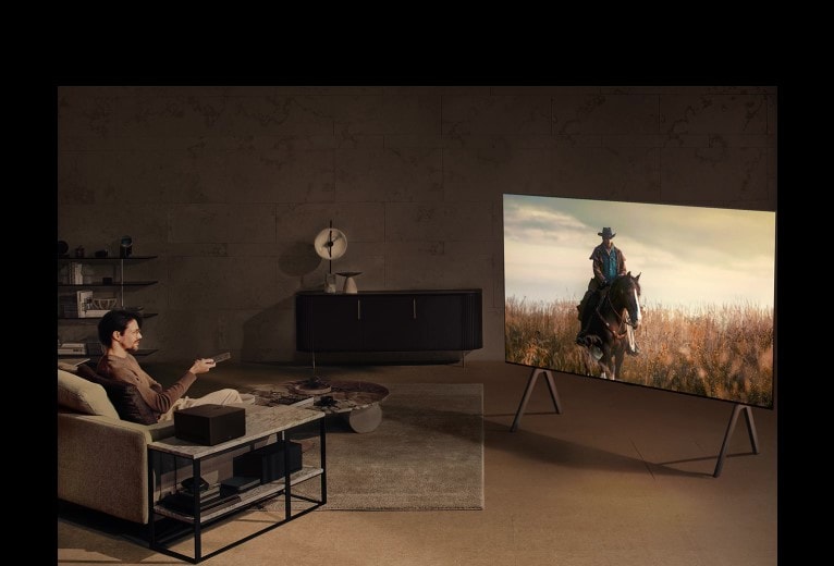 Egy férfi este cowboyfilmet néz a kutyájával egy hangulatos nappaliban. Nincsenek kábelek a falra szerelt LG OLED TV körül, a vezeték nélküli Connect Box pedig jól elfér egy oldalsó asztalon. 