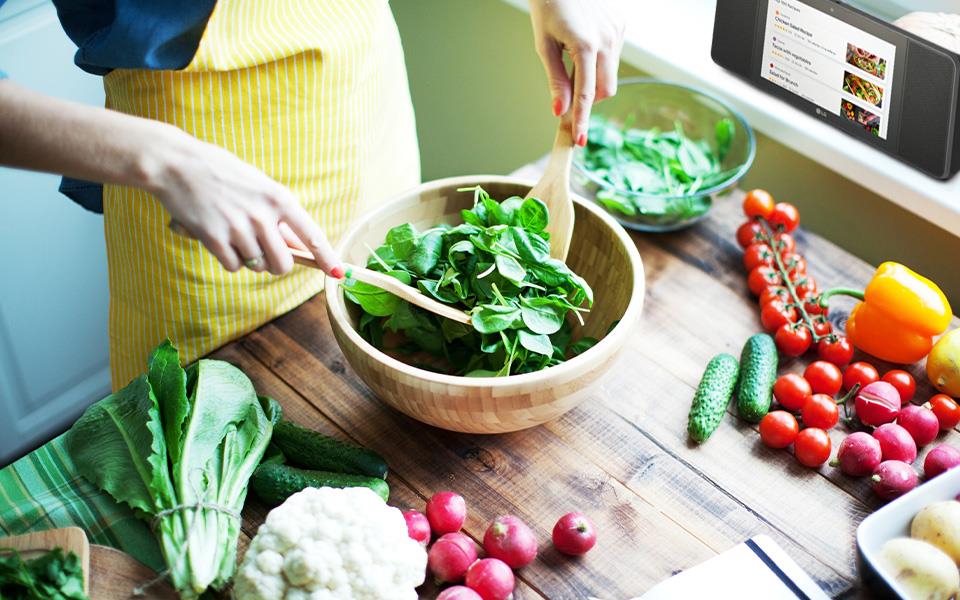 A person tossing a salad in a bowl while listening to a recipe on their LG speaker