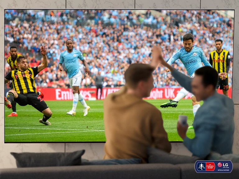 Dua orang bersorak sambil menonton pertandingan sepak bola pada NanoCell TV di ruang santai.