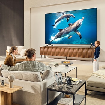 A family sits on a couch facing a LG QNED TV mounted on a wall above a LG Soundbar with a little girl pointing to a screen depicting two dolphins. The title talks about how every action is more thrilling on a massive screen.