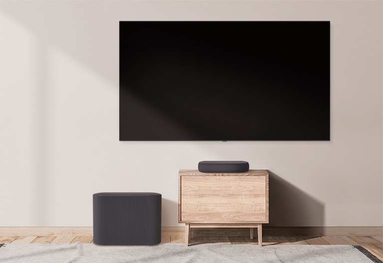 There is a bulky soundbar placed on a wide cabinet and a subwoofer placed on the left. The scene changes and a compact soundbar appears on a smaller cabinet and a subwoofer placed on the left.