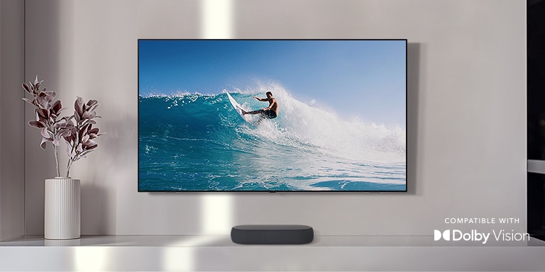 TV is on the wall. TV shows a man surfing on big wave. LG Soundbar is right below TV on a white shelf. There is a vase with a flower right next to the soundbar.Dolby Vision logo placed on right bottom corner.