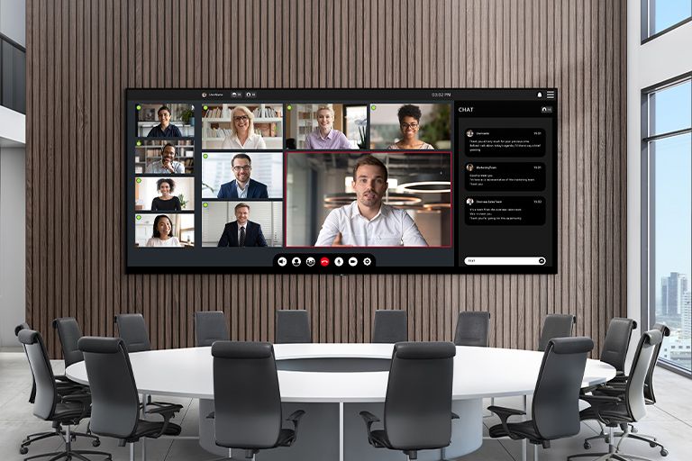 A wide LED screen is installed on the wall of a meeting room with a large round table. The expansive LED screen shows an ongoing video conference.