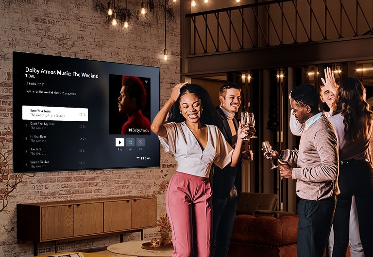 Four people dancing and enjoying drinks at a house party while music is playing on a large, wall-mounted LG QNED Mini LED TV.
