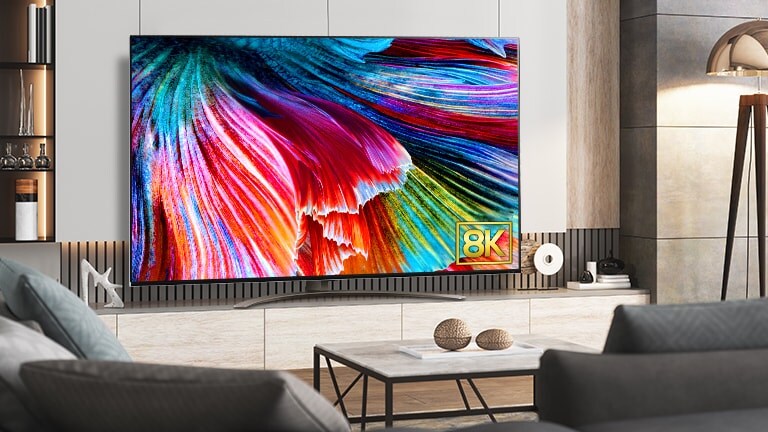 QNED99 stood against a wall in a light interior with sofas and a coffee table in front. The screen shows a close-up image of multi-colored petals and the 8K logo.