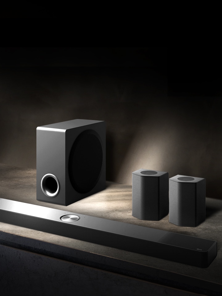 La LG Soundbar, gli altoparlanti posteriori e il subwoofer sono collocati in una prospettiva angolata su uno scaffale di legno marrone in una stanza nera, avvolta nell’oscurità con la luce che illumina solo il sistema audio.