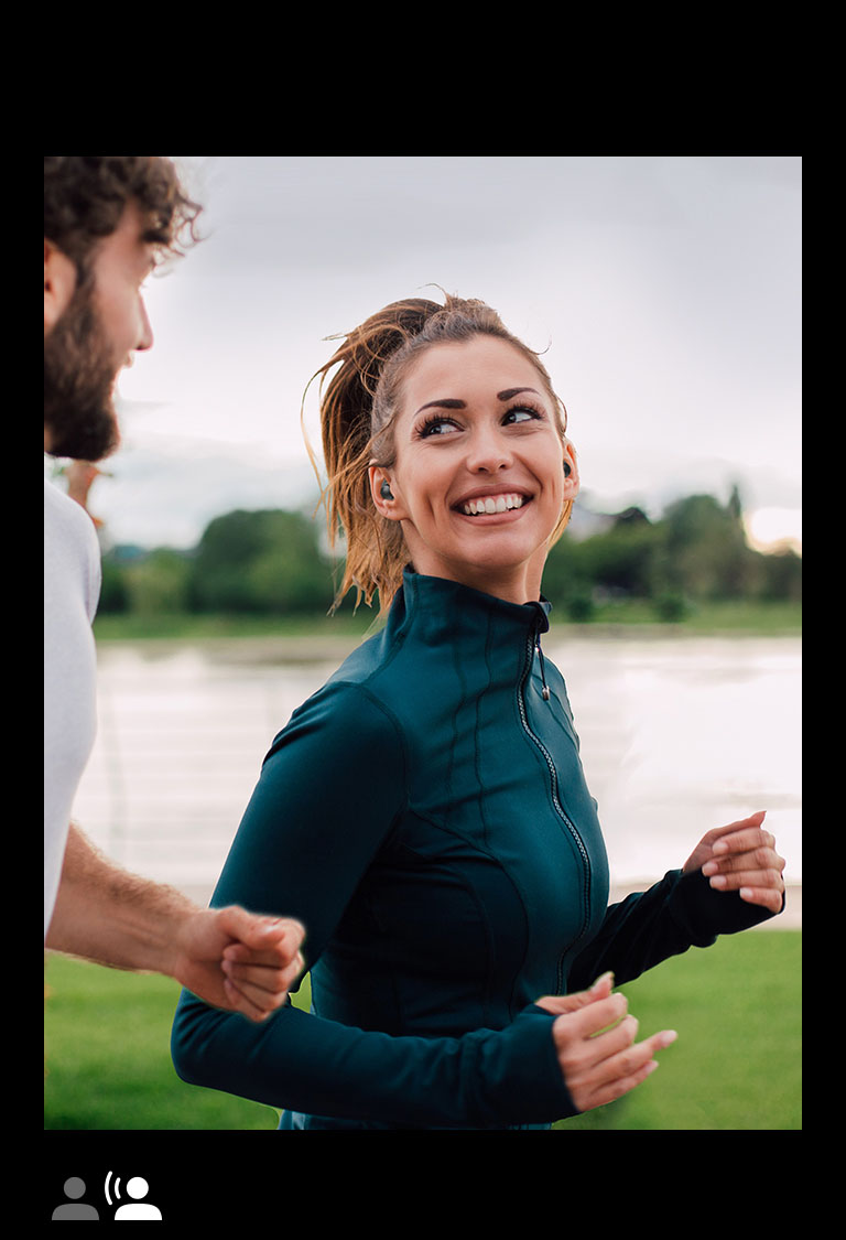 Un uomo e una donna corrono in un parco indossando le cuffie TONE Free Fit.