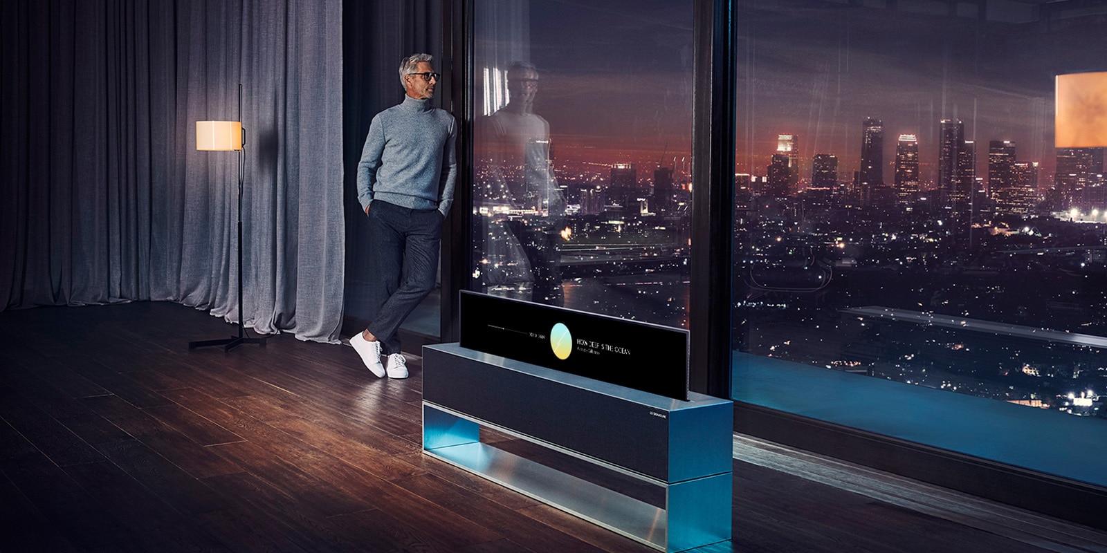 Un uomo si sporge da una finestra per ammirare il paesaggio notturno della città. Di fianco a lui c'è un LG SIGNATURE Rollale OLED TV in modalità Line View.