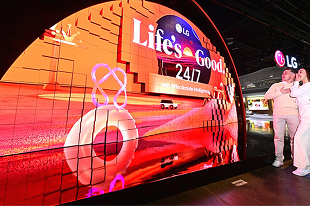 Visitatori osservano il display LED curvo di LG che mostra lo slogan “Life's Good 24/7” durante una fiera tecnologica.
