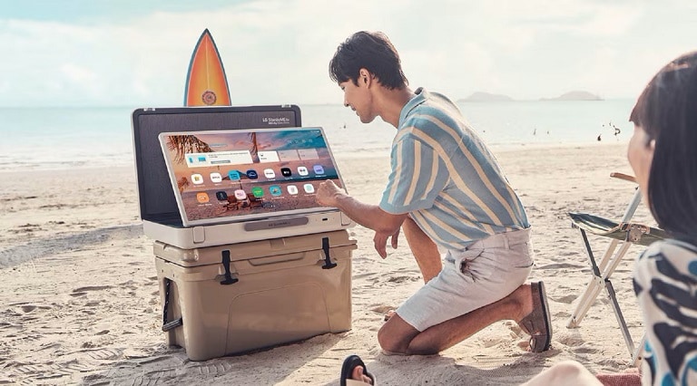 Un uomo che installa un dispositivo portatile LG su un frigorifero da spiaggia, con una tavola da surf sullo sfondo. Il dispositivo mostra un'interfaccia utente con varie app.