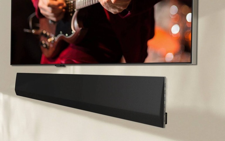 Una soundbar LG posizionata sotto un TV che riproduce l’immagine di una persona che suona una chitarra elettrica. La messa a fuoco è sulle mani e sulla chitarra, mentre uno sfondo caldo e sfocato crea un’atmosfera accogliente. La composizione sottolinea l’elevata qualità del sistema d’intrattenimento.