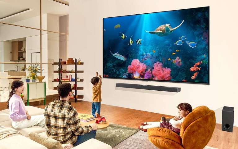 Una famiglia in un soggiorno che guarda una scena sottomarina con coralli e creature marine dai colori accessi su un TV LG montato a parete. Sotto il TV LG si trovano una soundbar e un subwoofer LG che migliorano l’esperienza visiva all’interno del soggiorno spazioso e moderno.