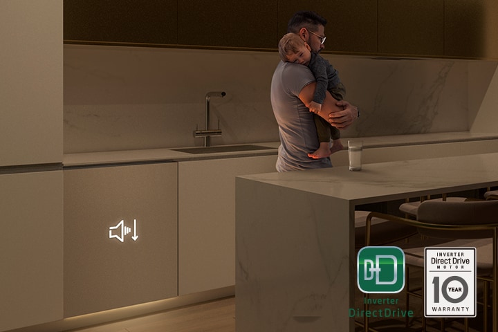 Мужчина держит на руках спящего ребенка на тускло освещенной кухне, а на заднем плане тихо работает отдельно стоящая посудомоечная машина.