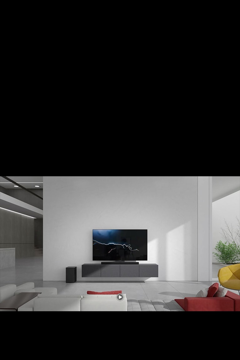 Саундбар размещен в гостиной на серой тумбе с телевизором. На полу слева расположен черный беспроводной сабвуфер, а с правой стороны изображения падает солнечный свет. Перед телевизором и саундбаром стоит бело-красный длинный диван.