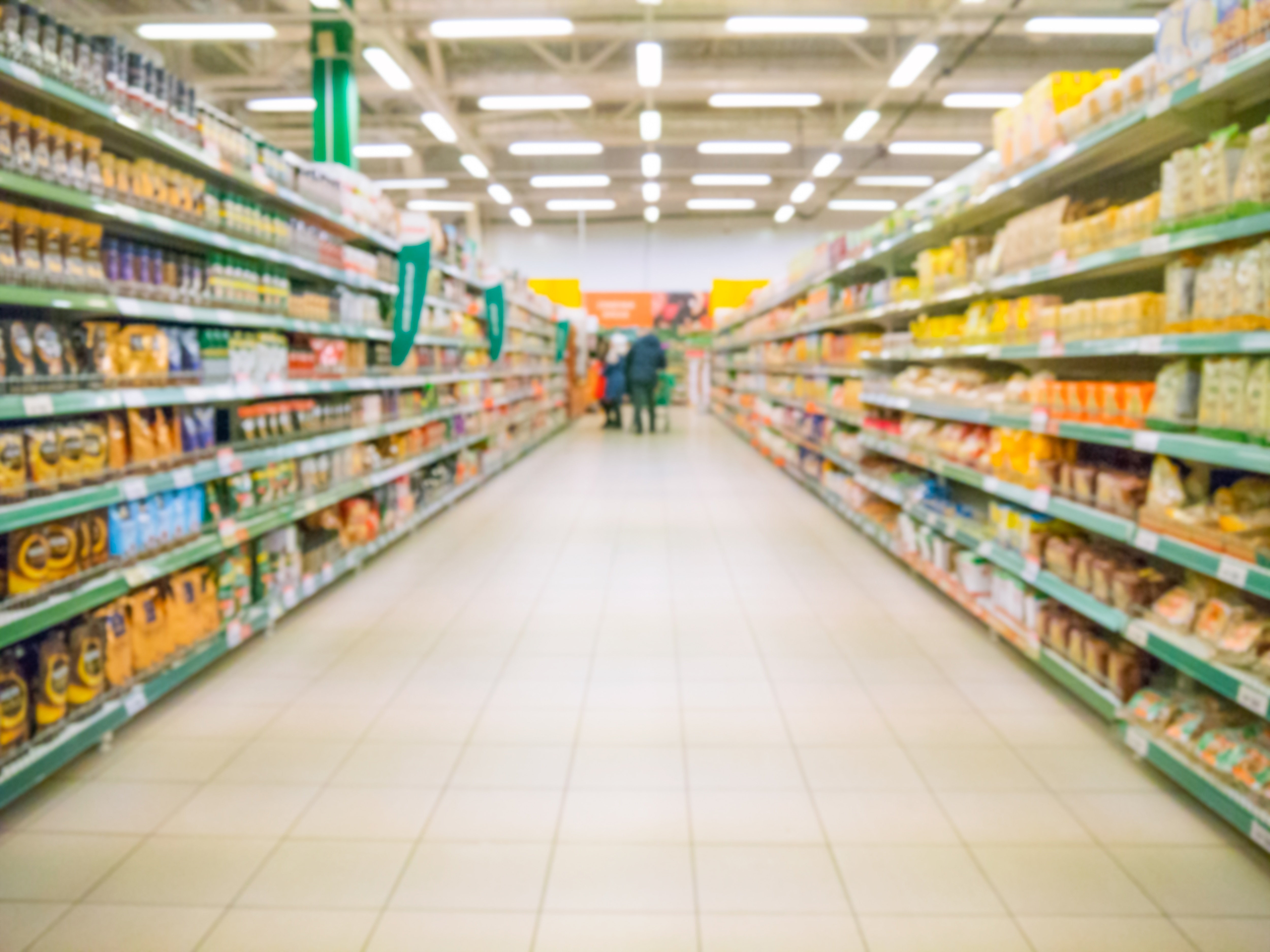 Pasillo de supermercado con estantes llenos de productos, enfoque en el centro donde se ven personas caminando.