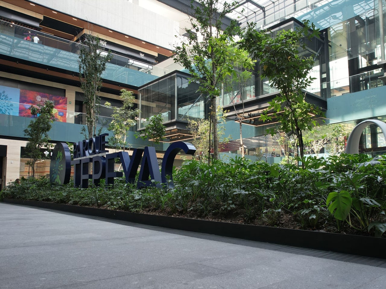 Moderno centro comercial con áreas verdes y un letrero grande en el centro, rodeado de edificios de vidrio y metal.