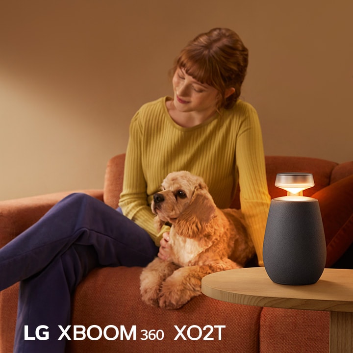 Una mujer sentada en el sofá con su perro y escuchando música con LG XBOOM 360 XO2T.
