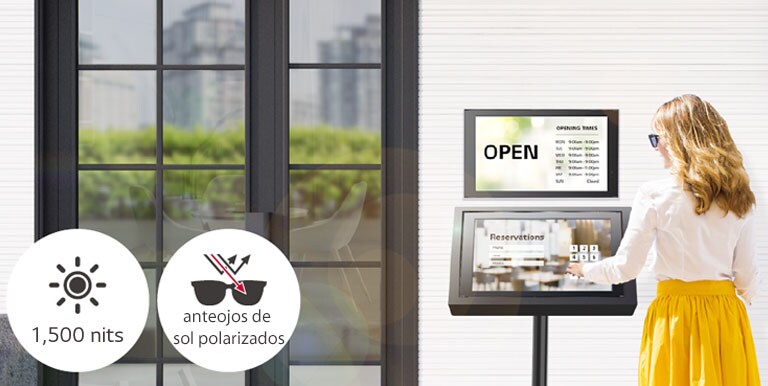 Se instala un 22XE1J en la pared a la entrada del edificio con otro soporte 22XE1J debajo para fines de reserva. La dama con gafas de sol está usando la pantalla 22XE1J con una superposición táctil para hacer una reserva. La pantalla permanece visible incluso bajo la luz solar directa.