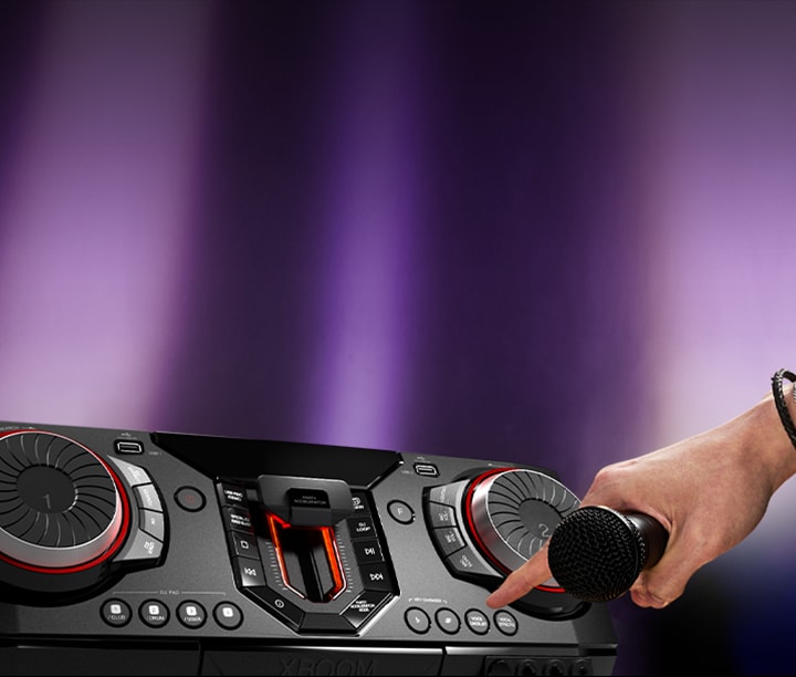 La imagen muestra un fondo borroso con focos de luz violeta. En el lado izquierdo de la imagen, hay un primer plano de la vista lateral del receptor, con una mano que sostiene un micrófono. El dedo índice está extendido para presionar el botón de karaoke