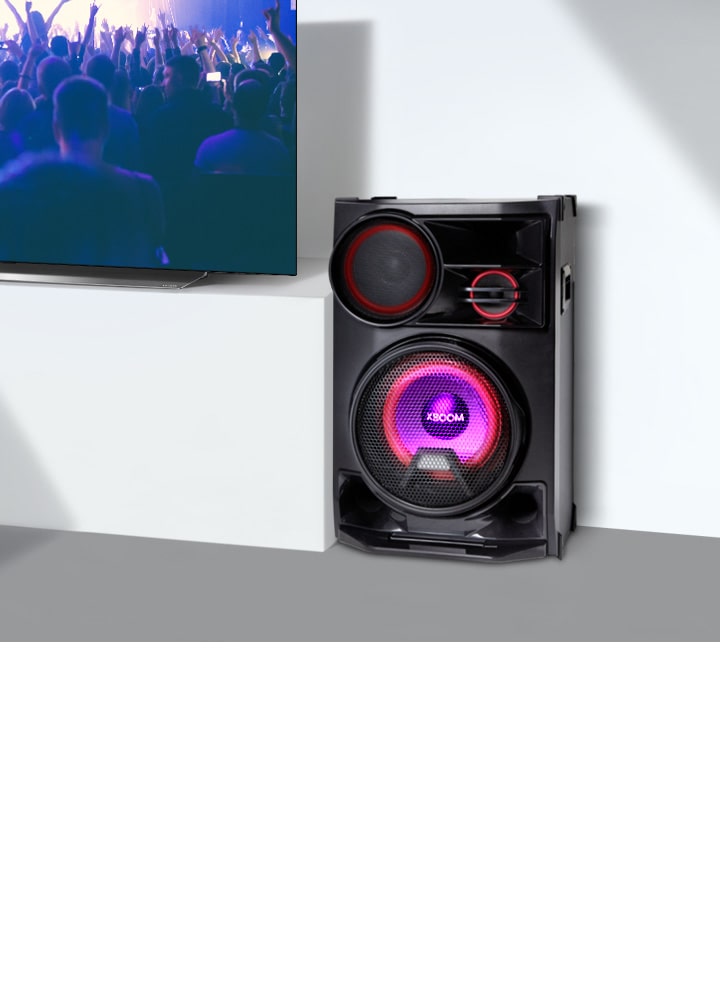 La imagen muestra un primer plano de una mesa larga y blanca para televisión. A la izquierda, un televisor LG muestra una escena de un concierto. En el lado derecho del televisor, hay un altavoz colocado sobre la mesa.