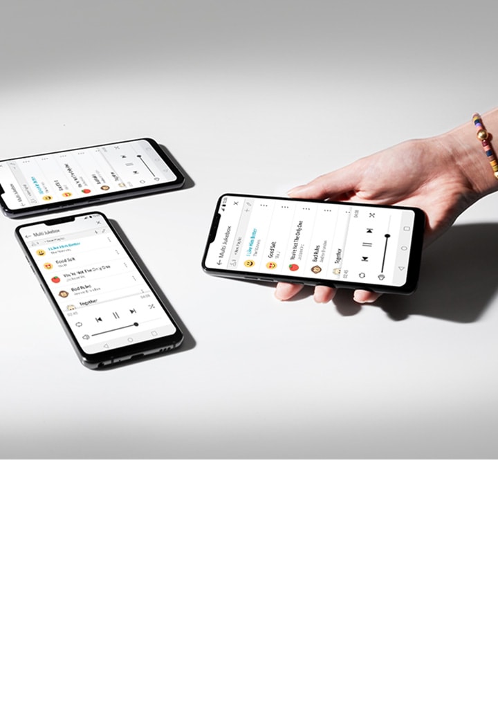 La imagen muestra una superficie blanca con luz que proviene del lado izquierdo. Hay tres teléfonos inteligentes colocados sobre la superficie blanca y una mano sostiene uno de ellos. Todos los teléfonos inteligentes muestran la lista de reproducción de Multi Jukebox.