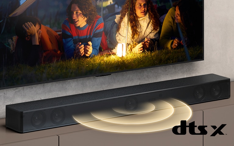 LG TV está en la pared, en la pantalla muestra 2 parejas tiradas en el césped. Frente a ellos, hay una lámpara. La barra de sonido LG está debajo del televisor LG. El gráfico de sonido sale del frente de la barra de sonido. El logotipo de DTS Virtual:X se muestra en la esquina inferior derecha de la imagen.