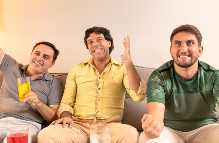 Tres amigos sentados en un sofá, todos levantando los brazos como si estuvieran animando en el partido de fútbol. Todos sonriendo.